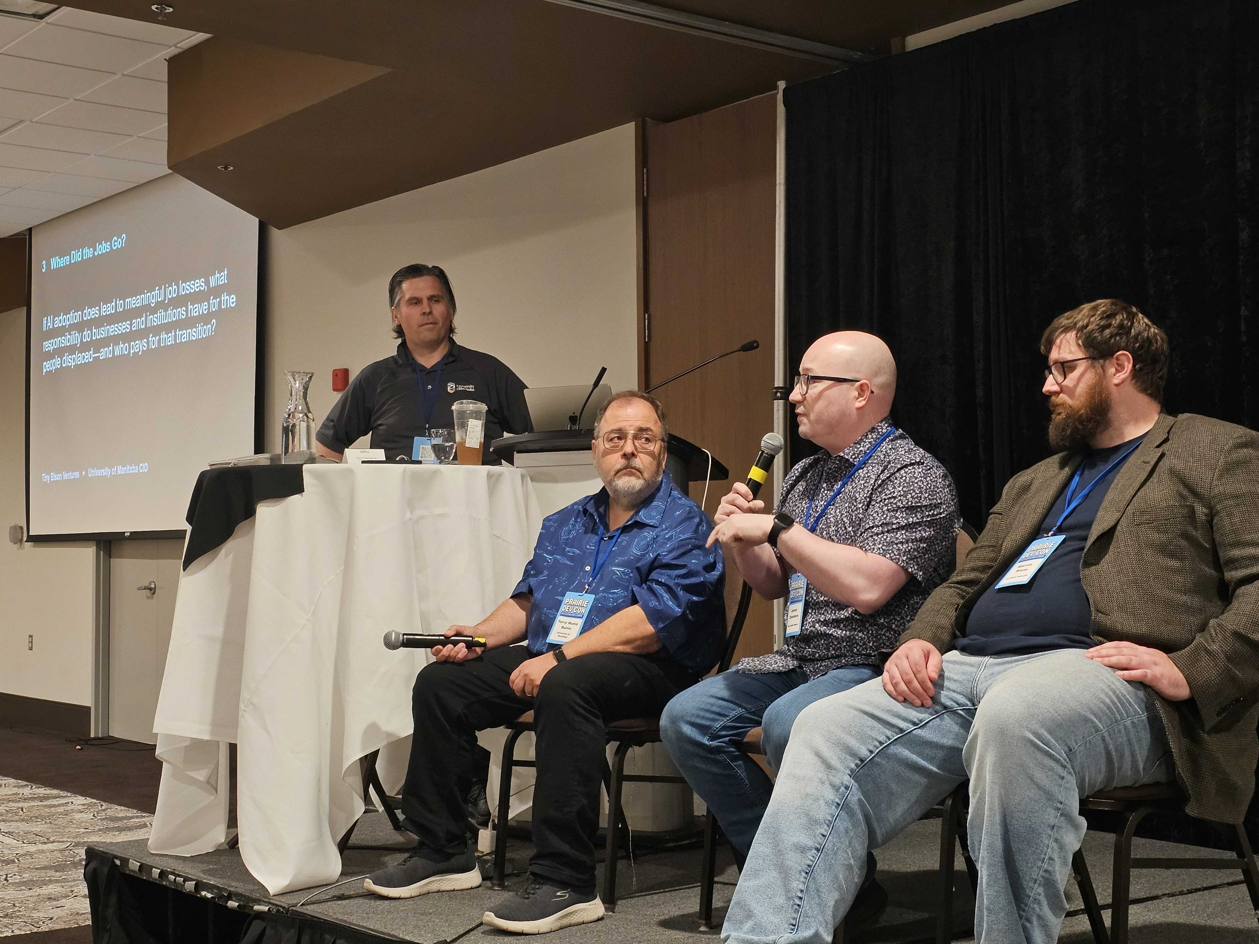 Panel discussion on stage with three seated speakers and a moderator at a podium. One speaker is holding a microphone while the others listen, and a projected slide behind them asks about AI adoption, job losses, and responsibility for displaced workers.